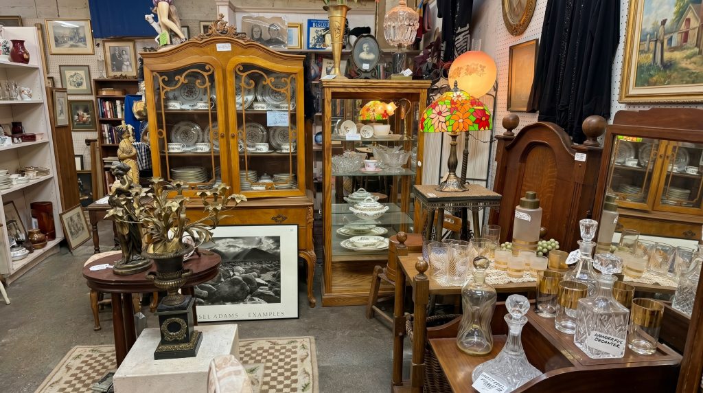 A curated display of traditional fine antiques featuring a large wood china cabinet with floral dinnerware, a stained glass floral lamp, and various crystal decanters and gold-rimmed glassware on a wooden bar cart.