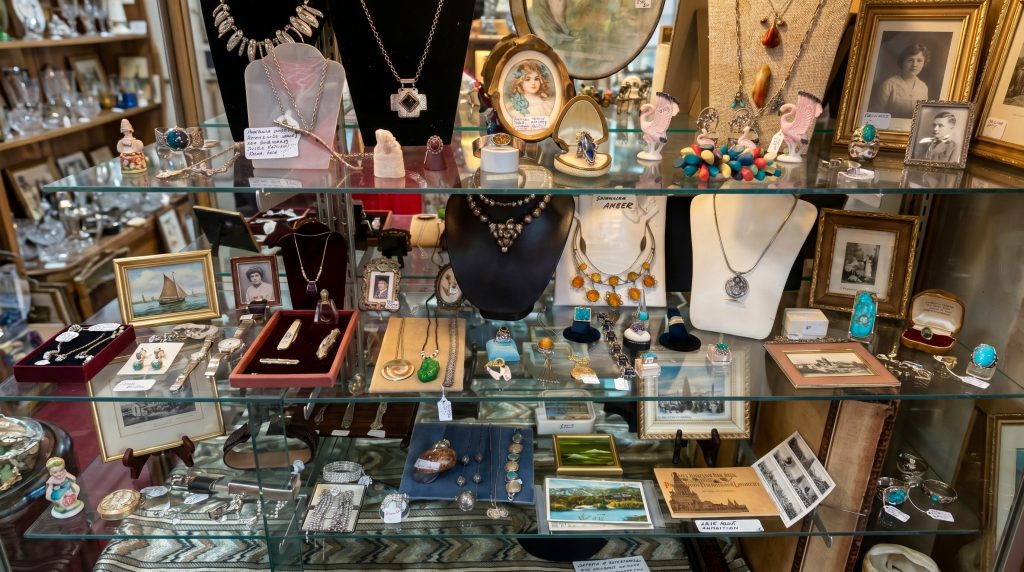 A curated glass display case featuring vintage estate jewelry, including silver necklaces, gemstone rings, turquoise bracelets, and antique cameo portraits in a brightly lit showcase.